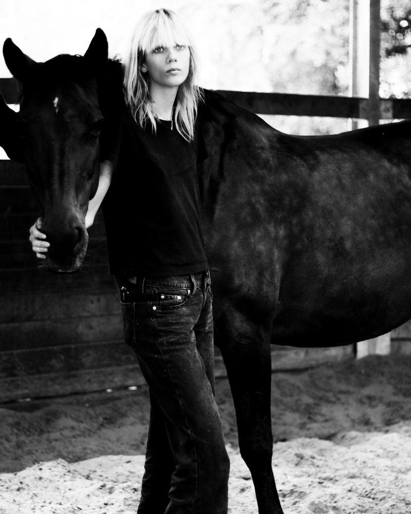 A woman stands next to a horse in a black and white photograph. The woman has light hair and is wearing dark clothes, the horse is dark.