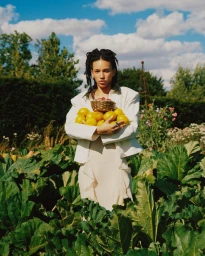 A woman holding a basket of fruit and lemons standing amidst lush garden foliage under a partly cloudy sky.