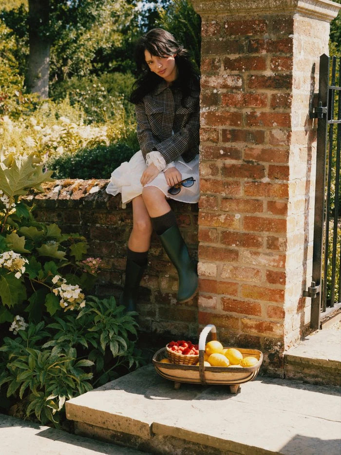 A woman in a plaid jacket and green wellington boots sits on a brick wall next to a basket of lemons.