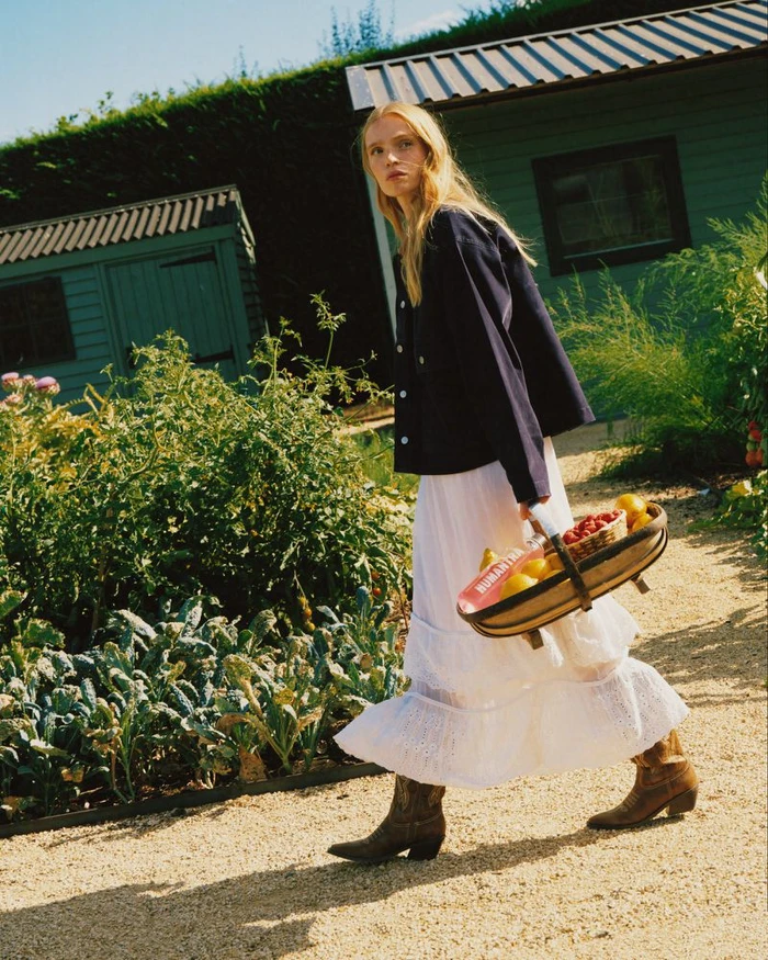 Blonde woman wearing a dark blue jacket and white skirt walking outdoors past green sheds carrying a basket of fruit
