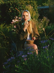 Woman in a silk headscarf and dark jacket seated outdoors among garden flowers holding a small woven basket