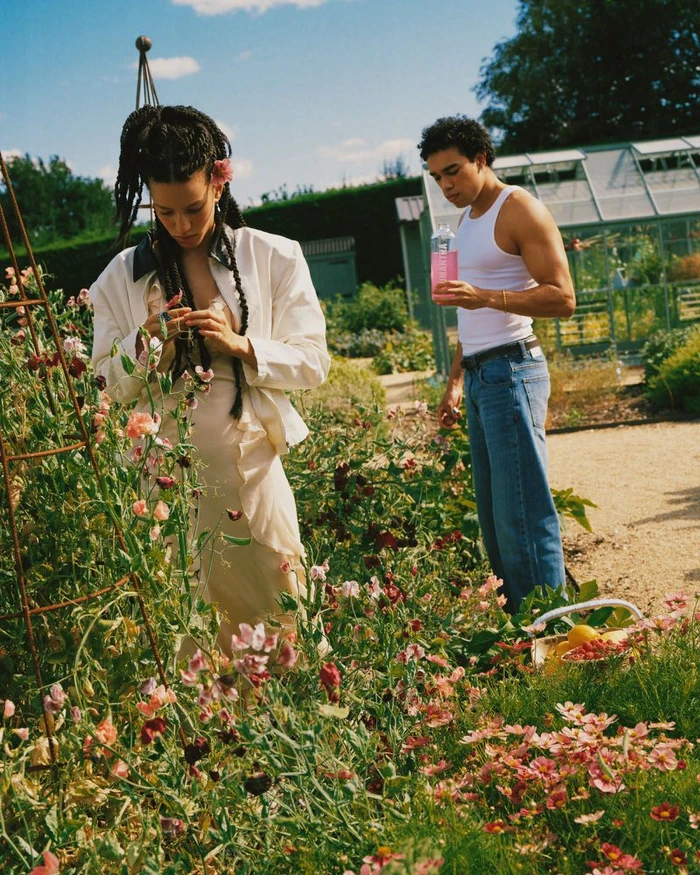 Woman in white jacket and slip dress standing among flowers, with a man holding a bottle nearby in a garden.
