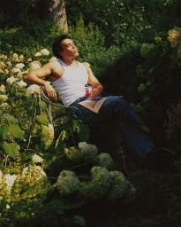 Young man in a white tank top and blue jeans seated on a bench outdoors surrounded by lush green foliage and flowers