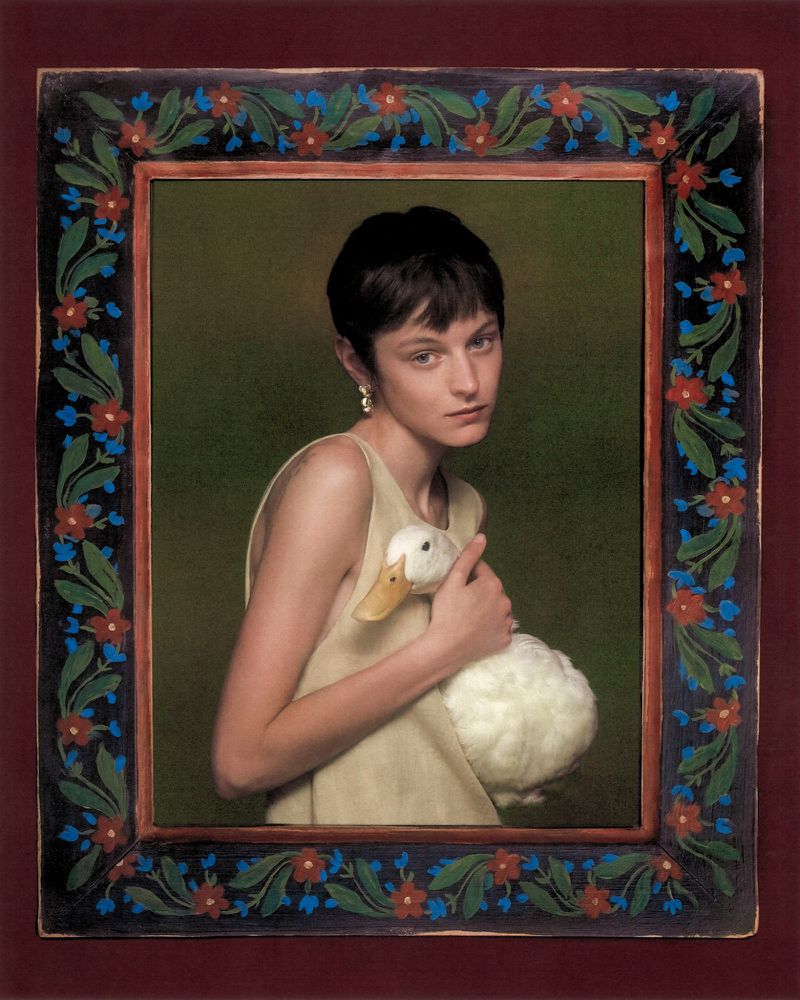 Portrait of Emma Corrin holding a white duck within an ornate painted floral frame for Harper's Bazaar UK.