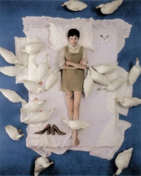 Emma Corrin lying flat on a bed surrounded by white ducks with a blue floor background for Harper's Bazaar UK.