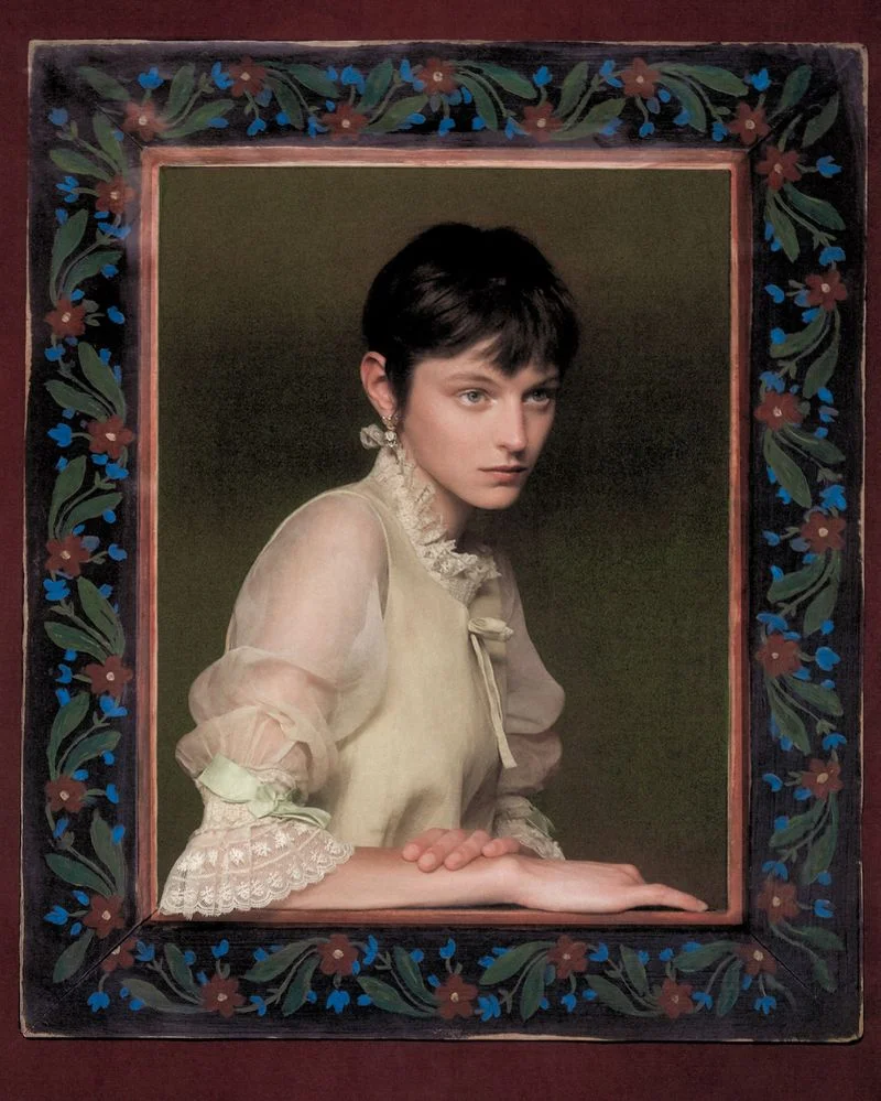 Portrait of Emma Corrin wearing a ruffled cream top framed by a painted floral border
