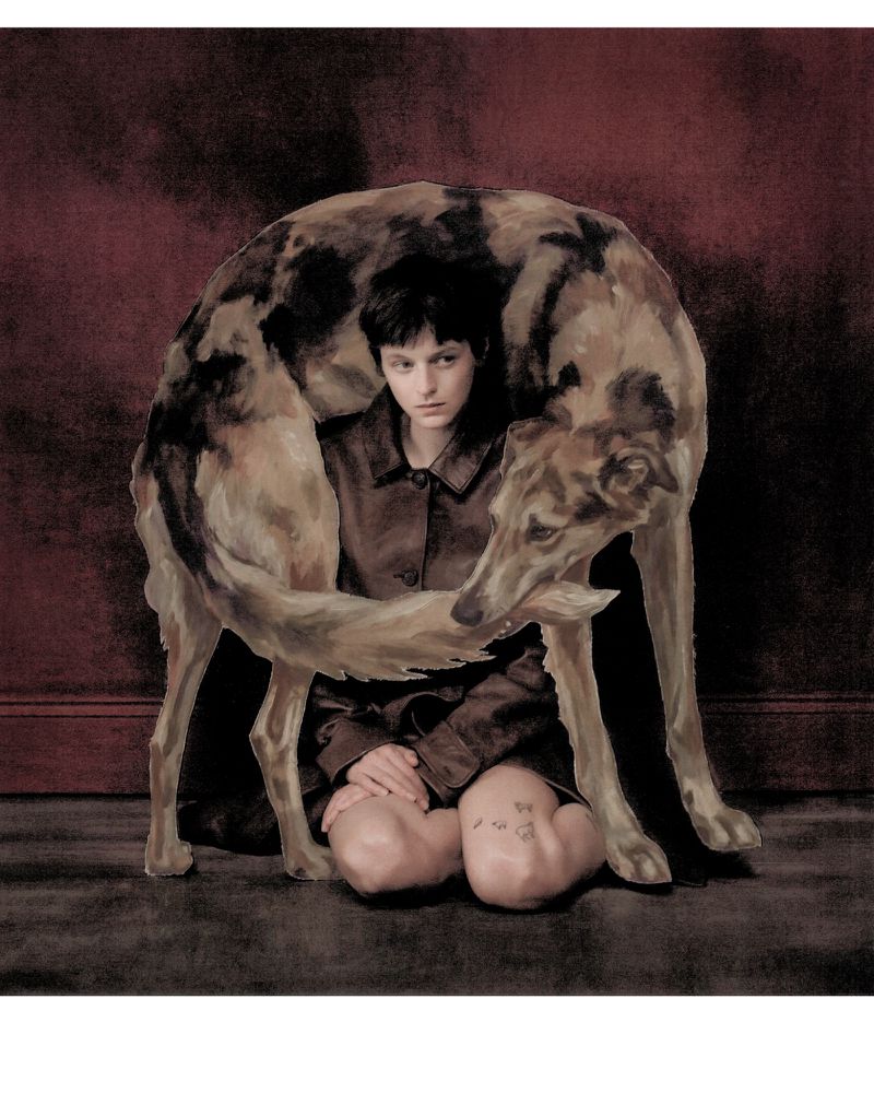 Emma Corrin kneeling on a dark floor framed by a large illustrated hound draped overhead for Harpers Bazaar UK
