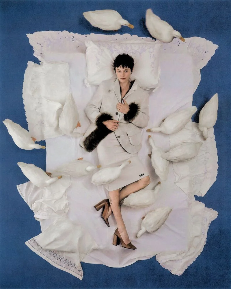 Emma Corrin lying on white bedding surrounded by white ducks on a blue surface for Harper's Bazaar UK.