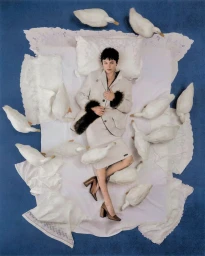 Emma Corrin lying on white bedding surrounded by white ducks on a blue surface for Harper's Bazaar UK.