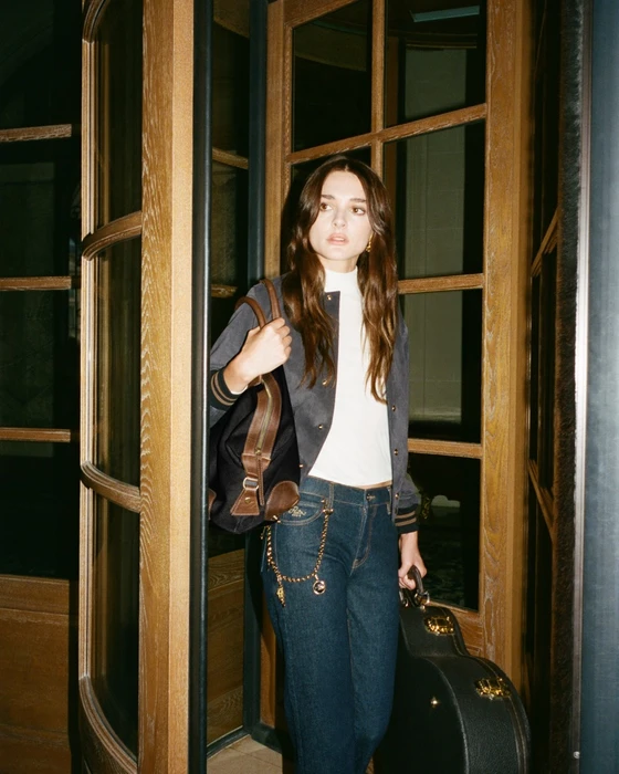 Charlotte Lawrence standing in a revolving wooden doorway holding a dark handbag and guitar case for FRAME