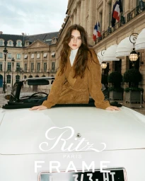 Charlotte Lawrence leaning on a white vintage convertible car outside a Parisian building in a brown suede coat