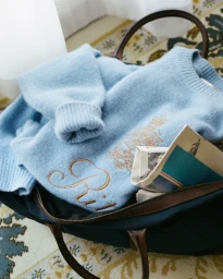 Light blue cashmere sweater with gold embroidery packed inside a dark canvas weekender bag with brown leather handles.