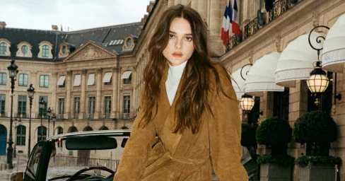 Charlotte Lawrence wearing a brown suede trench coat in Paris with a vintage car and historic building exterior.
