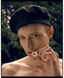 Close-up portrait of a young man wearing a black beret and holding a white daisy near his lips against a blurred green foliage background.