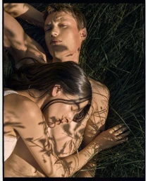 Two models lying in lush green grass with dappled sunlight filtering across them from overhead foliage.