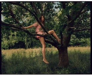 Model seated in the branches of a leafy tree wearing a pink crochet top and matching shorts in an outdoor meadow setting.