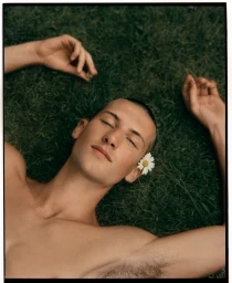 Close-up portrait of a man lying on green grass with a daisy tucked behind his ear