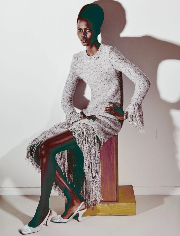 Model Ajah Angau Jok seated on a wooden block wearing a fringe dress and slingback heels against a white studio wall.