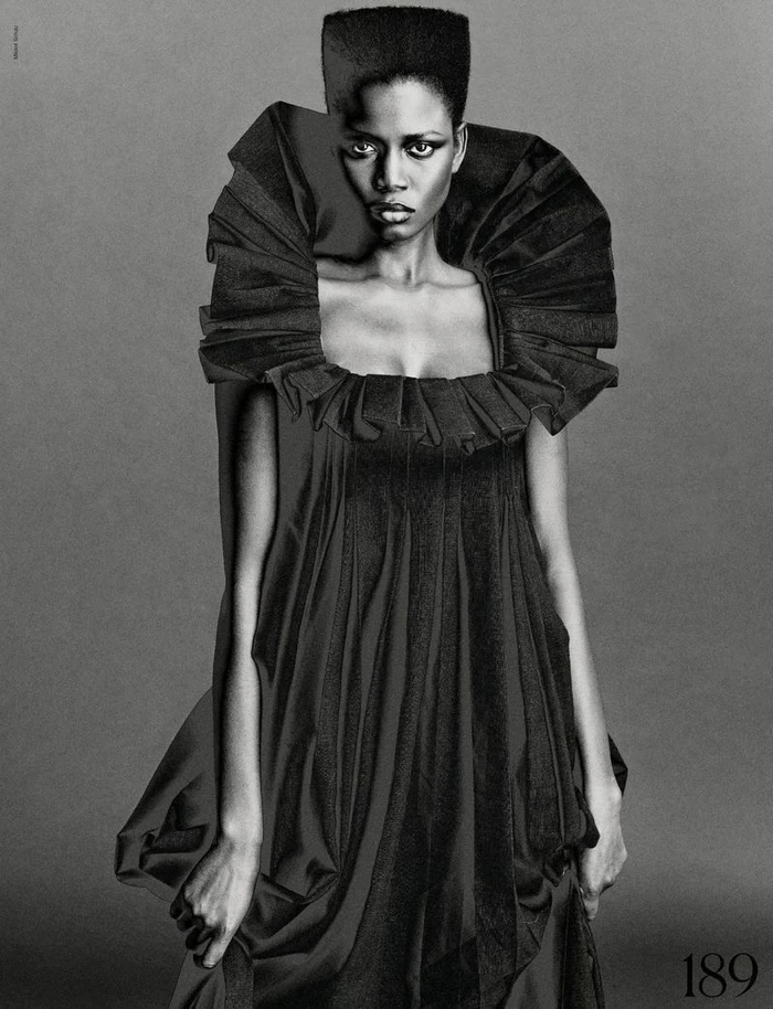Model Ajah Angau Jok in a black ruffled dress with a flat-top high hairstyle against a grey studio background.