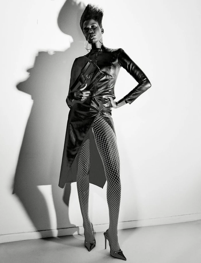 Model wearing dark leather clothing and fishnet tights standing against a bright white wall with a dramatic shadow