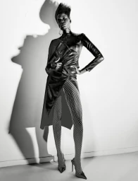 Model wearing dark leather clothing and fishnet tights standing against a bright white wall with a dramatic shadow