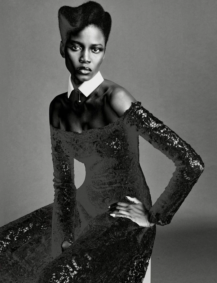 Model Ajah Angau Jok wearing a black lace off-shoulder dress and white collar against grey studio backdrop.