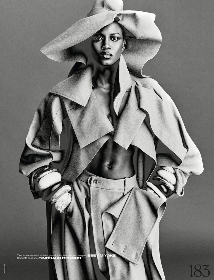Model Ajah Angau Jok wearing a wide-brimmed hat, structured coat, and bangles in a black and white editorial shoot by Mikael Schulz.