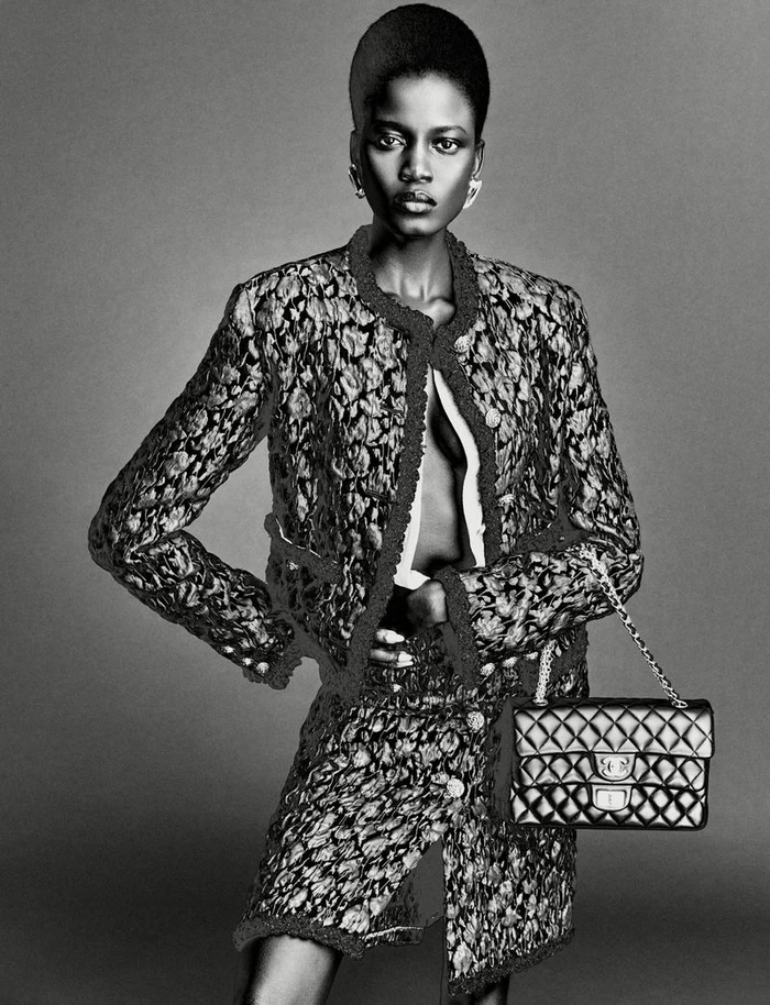 Black and white studio portrait of a model wearing a textured tweed jacket and skirt holding a quilted chain handbag.