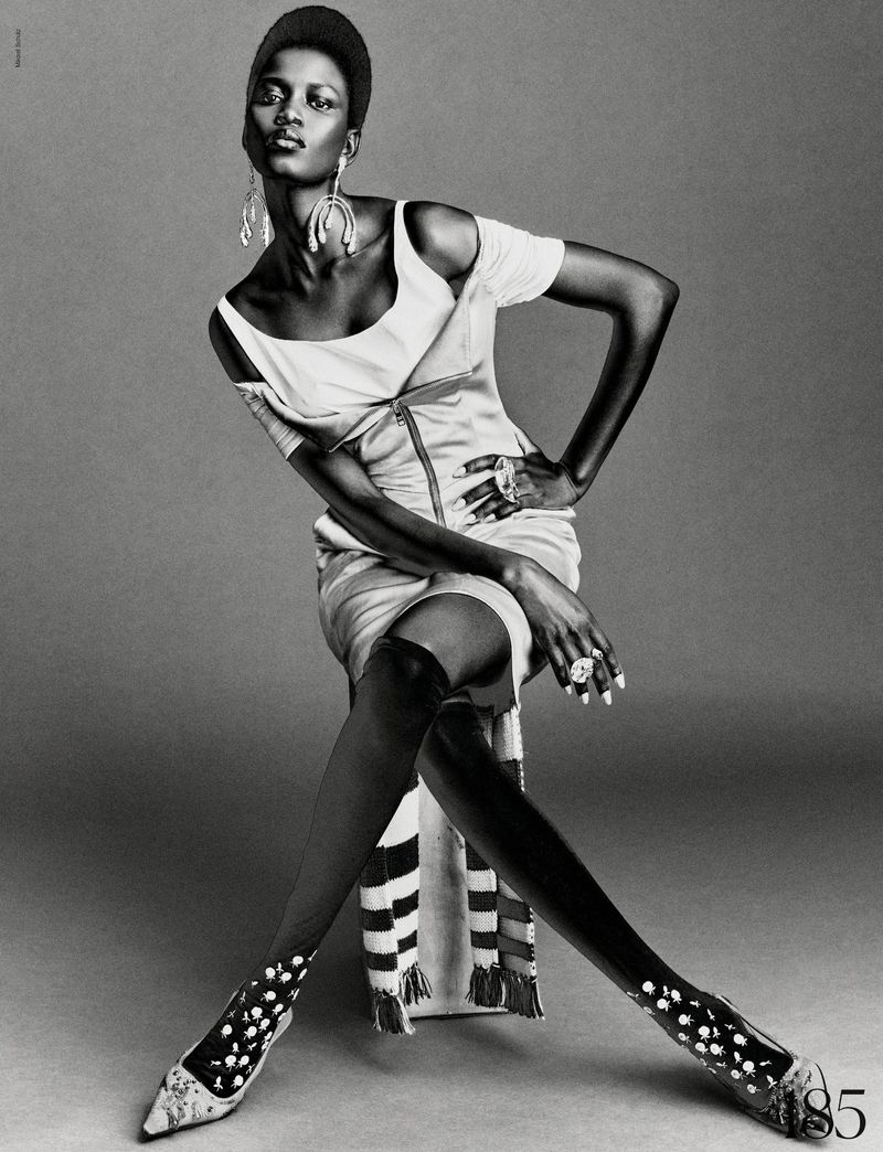 Model Ajah Angau Jok wearing a structured dress and tall boots seated against a grey backdrop for French Magazine