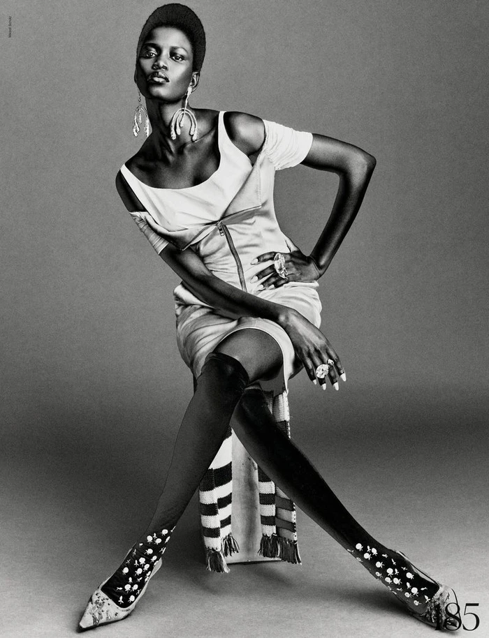 Model Ajah Angau Jok wearing a structured dress and tall boots seated against a grey backdrop for French Magazine