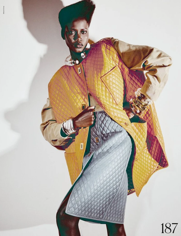 Model Ajah Angau Jok in a quilted yellow vest and grey skirt with bold accessories against a white studio background.