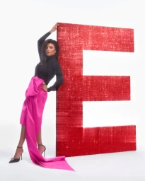 Eva Longoria leaning against a giant red glitter letter E, wearing a black turtleneck and bright pink satin skirt.