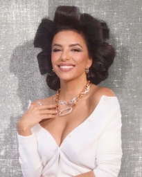 Eva Longoria smiling in a white cardigan with voluminous retro hair and layered gold jewelry against a sparkling textured background