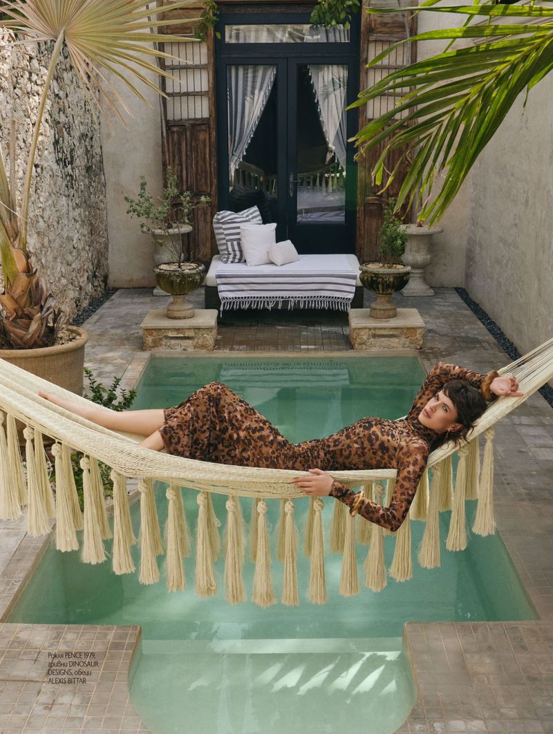 Woman in leopard print dress lounges in hammock above a pool, surrounded by stone walls and greenery. Sunny outdoor scene.