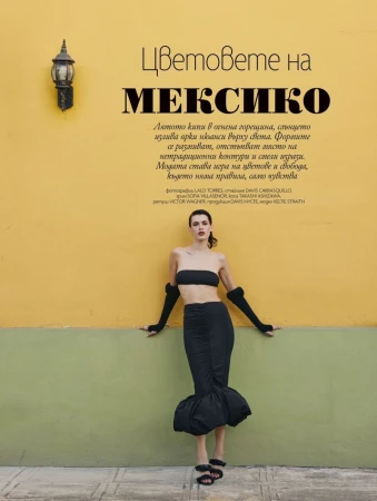 A woman in a black bandeau top and long black skirt poses against a two-tone yellow and green wall with a light fixture above.