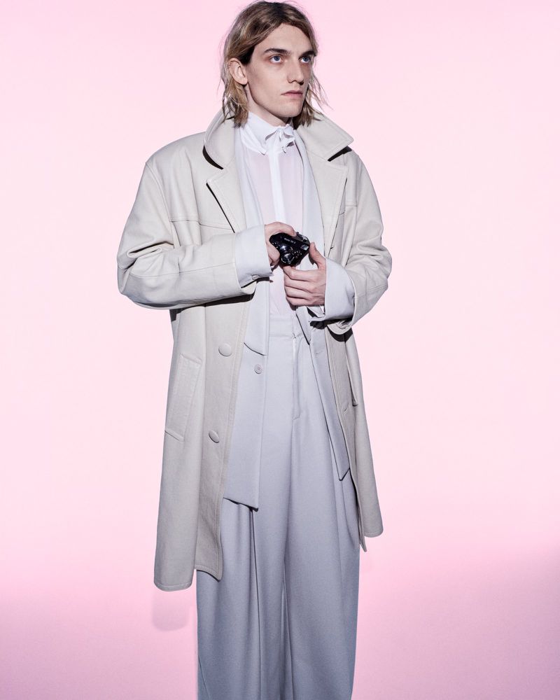 Male model in a cream long coat and wide-cut trousers holding a camera against a soft pink seamless background