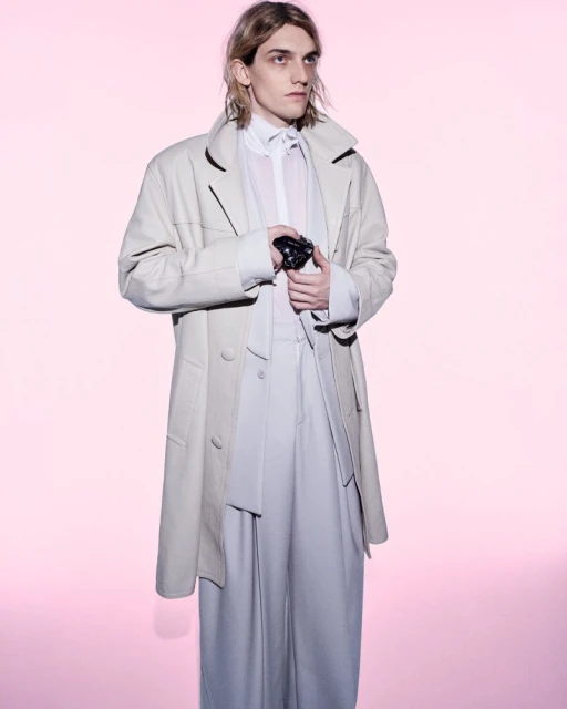 Male model in a cream long coat and wide-cut trousers holding a camera against a soft pink seamless background