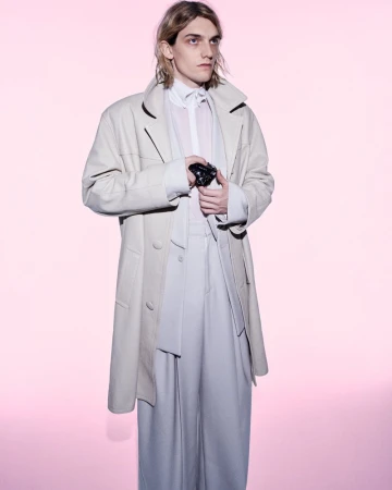 Male model in a cream long coat and wide-cut trousers holding a camera against a soft pink seamless background