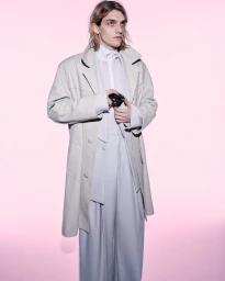Male model in a cream long coat and wide-cut trousers holding a camera against a soft pink seamless background
