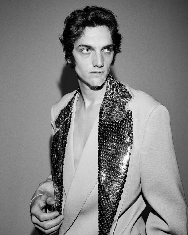 Monochrome portrait of a male model wearing a light-colored suit jacket with heavily textured sequined lapels against a plain background