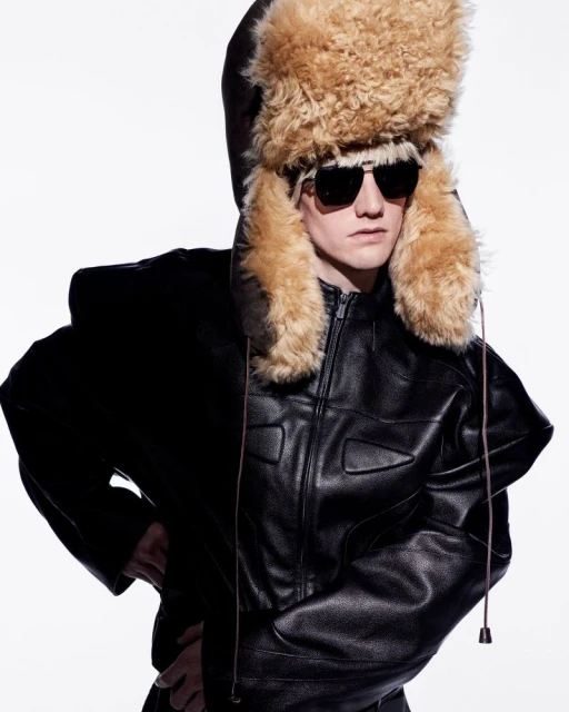 Model wearing a large furry trapper hat, dark sunglasses, and a black leather jacket against a plain white background.