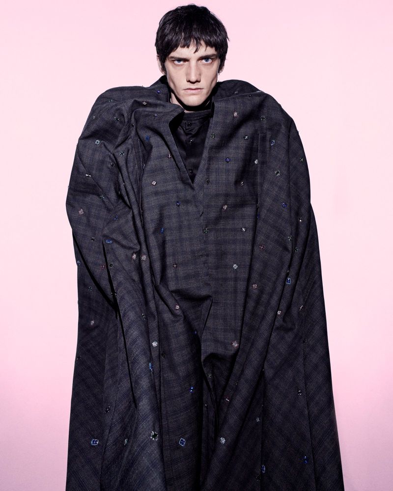 Male model in a dark patterned cloak standing against a soft pink gradient background for The Usual Suspect editorial.