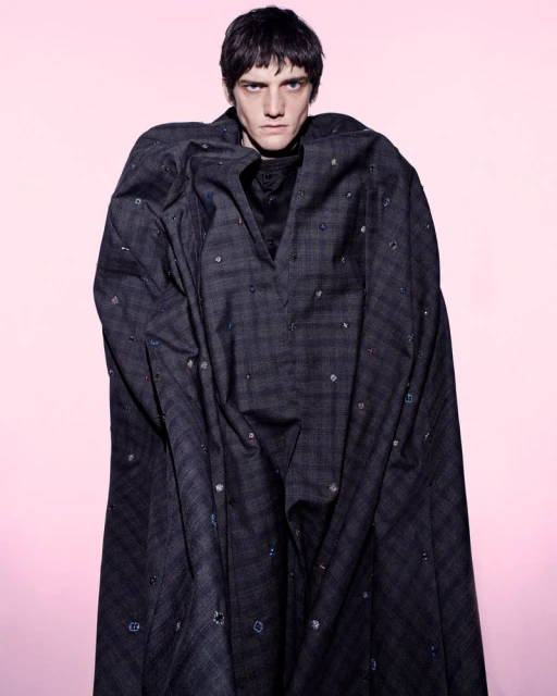Male model in a dark patterned cloak standing against a soft pink gradient background for The Usual Suspect editorial.