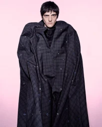 Male model in a dark patterned cloak standing against a soft pink gradient background for The Usual Suspect editorial.
