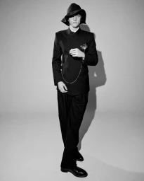 Monochrome portrait of a male model wearing a dark suit, tailored trousers, and a wide-brimmed hat in a studio setting.