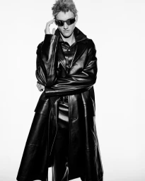 Model wearing dark sunglasses and a long black leather coat against a white background