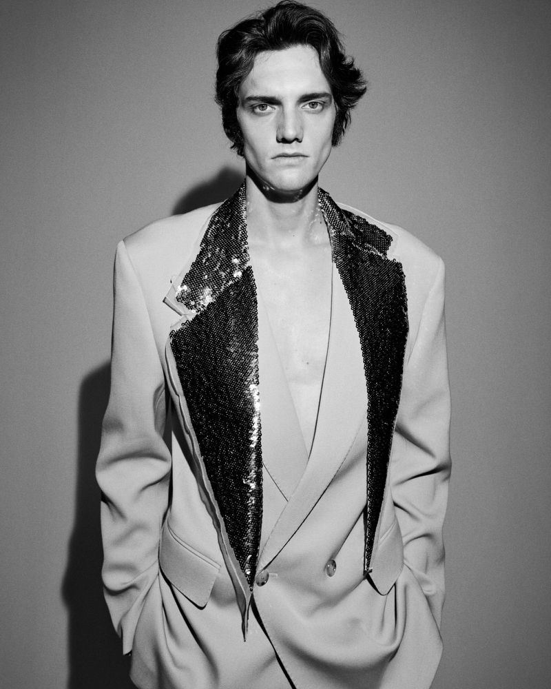 Portrait of a male model wearing a tailored blazer with sequin lapels against a plain grey studio backdrop.
