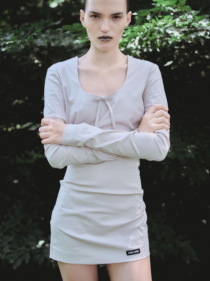 Model standing with arms crossed wearing a light grey long-sleeve dress against a blurred green foliage background