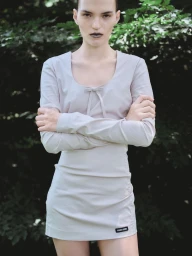 Model standing with arms crossed wearing a light grey long-sleeve dress against a blurred green foliage background
