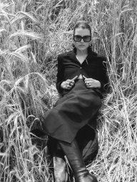 Model in black sunglasses and dark jacket seated amidst tall dry grass in black and white
